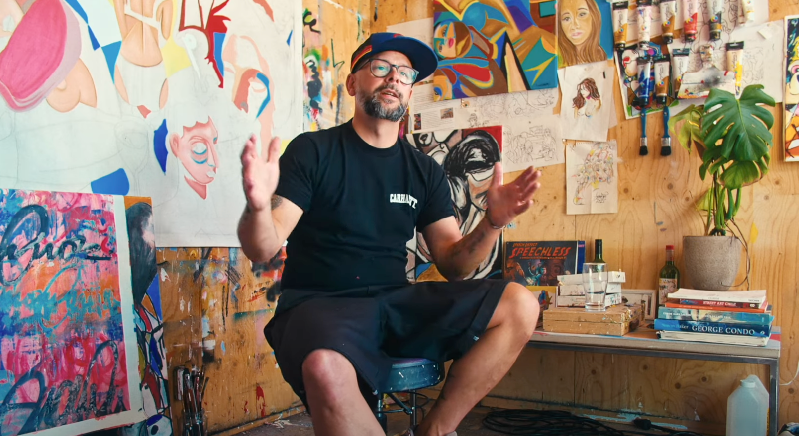 Man sitting in an artist's studio surrounded by artwork and art supplies.