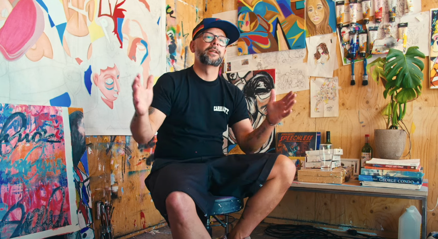 Man sitting in an artist's studio surrounded by artwork and art supplies.